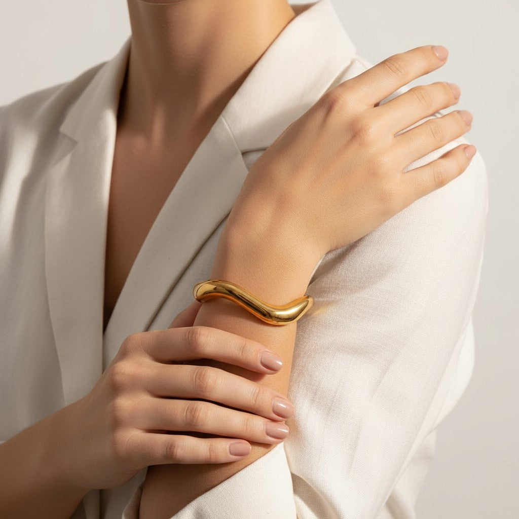 Lyza Jewels, LLC gold-toned 18k stainless steel wavy bangle cuff worn on a model in a white blazer.